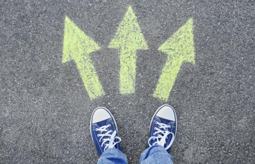 Image taken from above/point of view showing the legs of person a wearing blue jeans and a pair of canvas sneakers; the feet are aligned with arrows chalked on the pavement to indicate the concept of multiple possible paths.