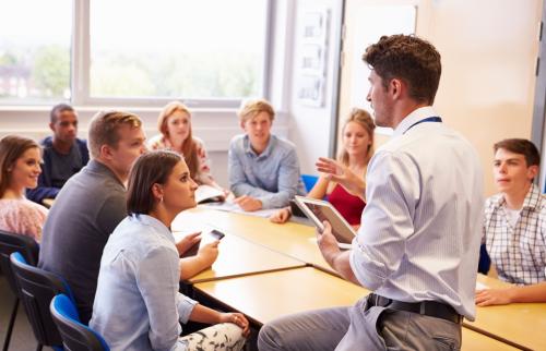 A professor speaking to a group of students seated around a table in a sunlit classroom